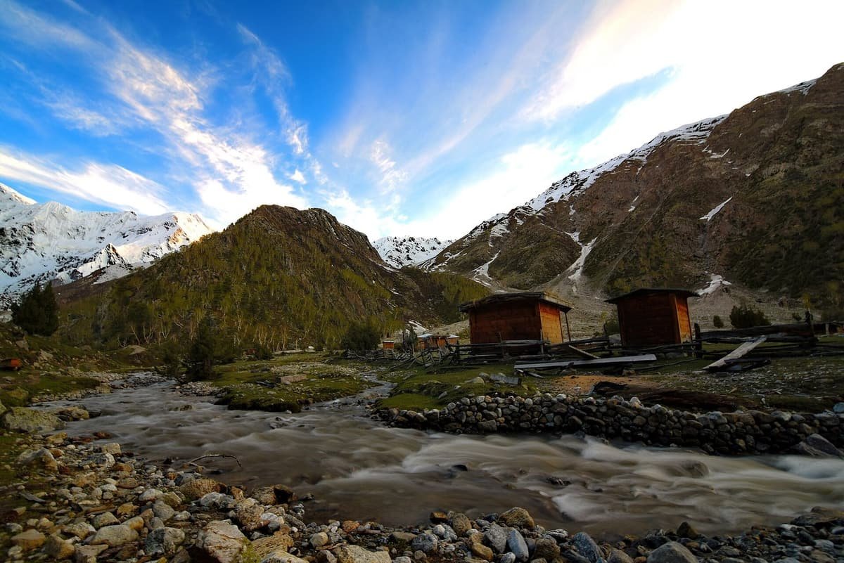 Wooden huts of Beyal Camp stand beside a rushing stream, with snowy peaks and green slopes under a blue sky.