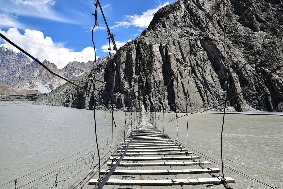 The empty Hussaini suspension bridge stretches over the grey Hunza River toward a sheer rock cliff and jagged peaks.