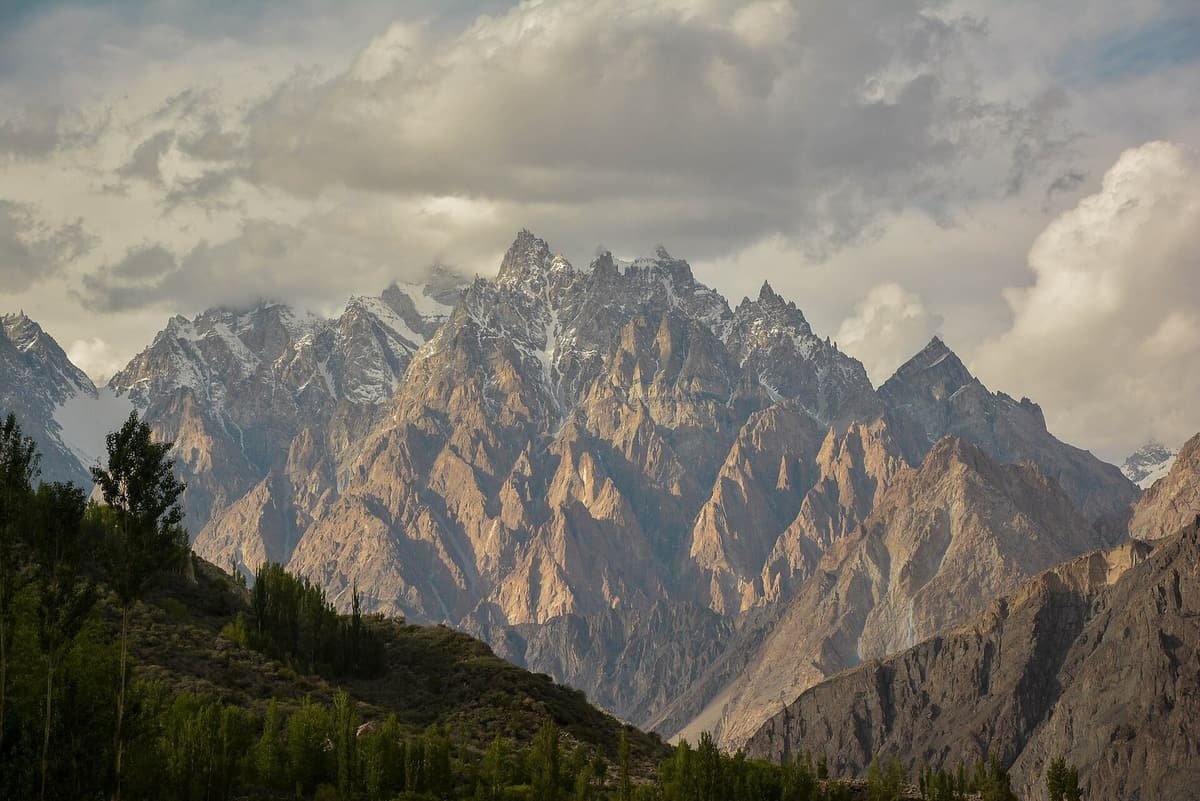 The jagged spires of the Passu Cones catch evening light under heavy clouds, with poplars on a dark hillside below.