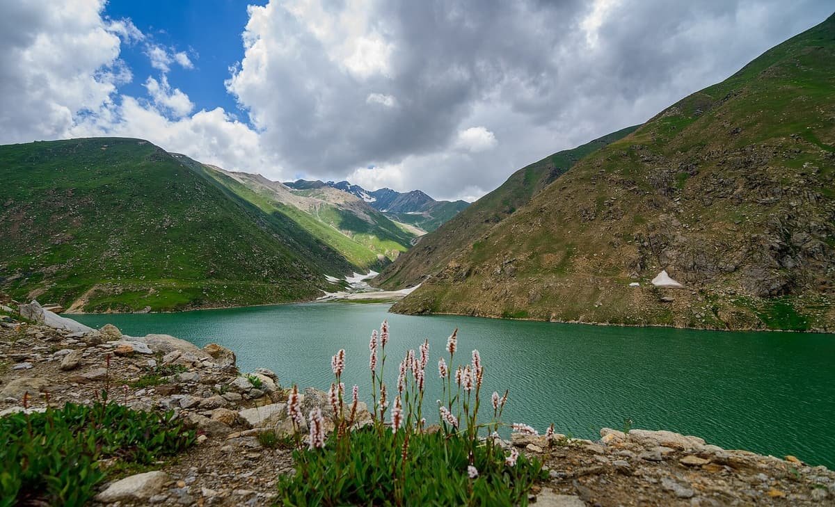 Jade-green Lulusar Lake sits between grassy mountain slopes under billowing clouds, with pink wildflowers on the rocky shore.