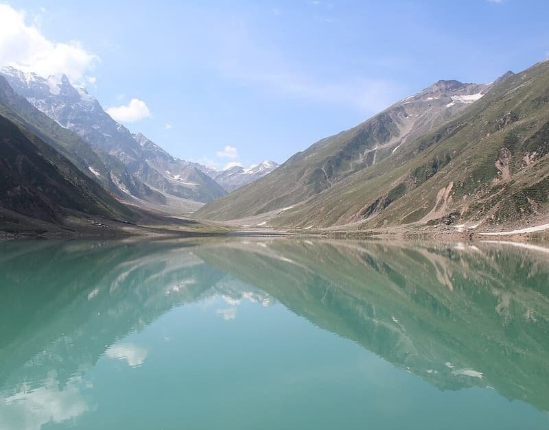 Lake Saiful Muluk lying still, mirroring green slopes and the snowy peaks above it