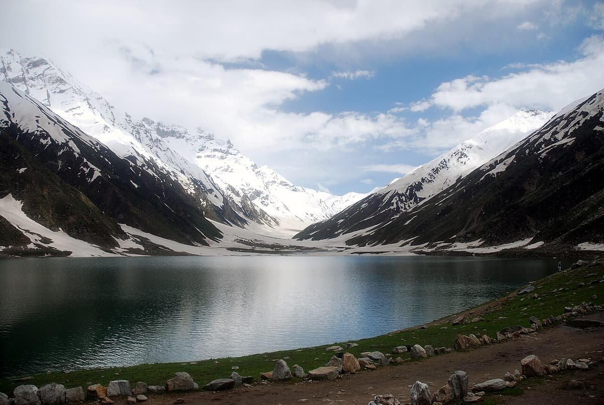 Lake Saiful Muluk lying still under snow-streaked mountains, with a stony path along its shore