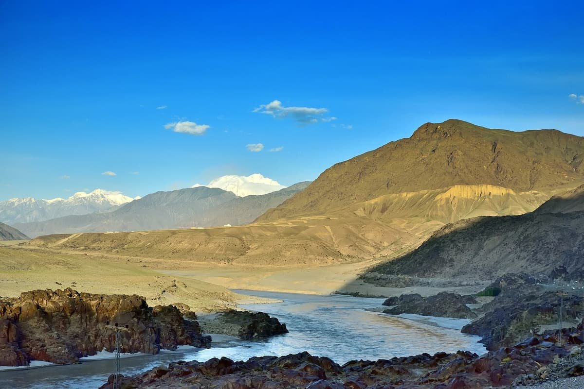 The Indus River winds between dark rocks and dry tawny hills near Chilas, with snowy peaks on the horizon.