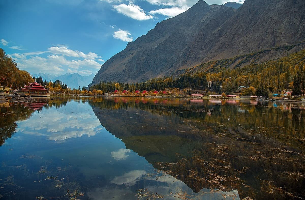 Lower Kachura Lake mirrors a steep mountain and autumn trees, with a red pagoda roof and small cottages on the far shore.