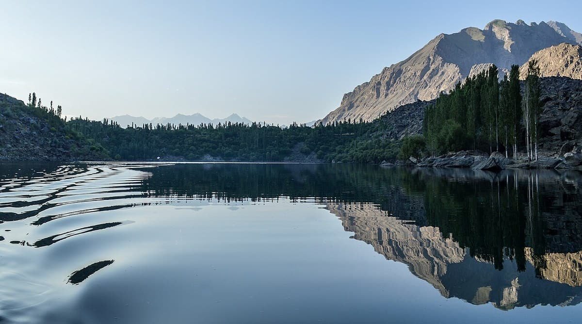 Still water of Upper Kachura Lake at dawn, reflecting a sunlit rocky peak and tall poplars