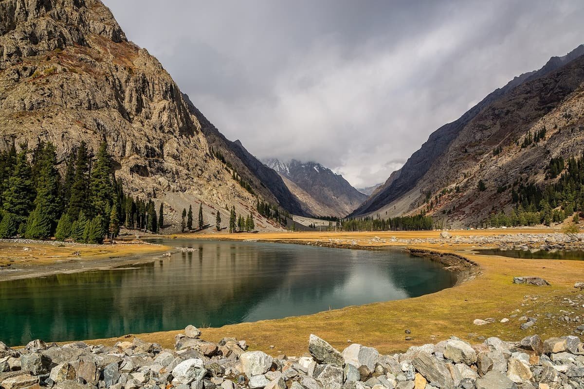 Turquoise Mahodand Lake curves through golden meadow grass toward a rocky valley under storm clouds, with pines along the shore.