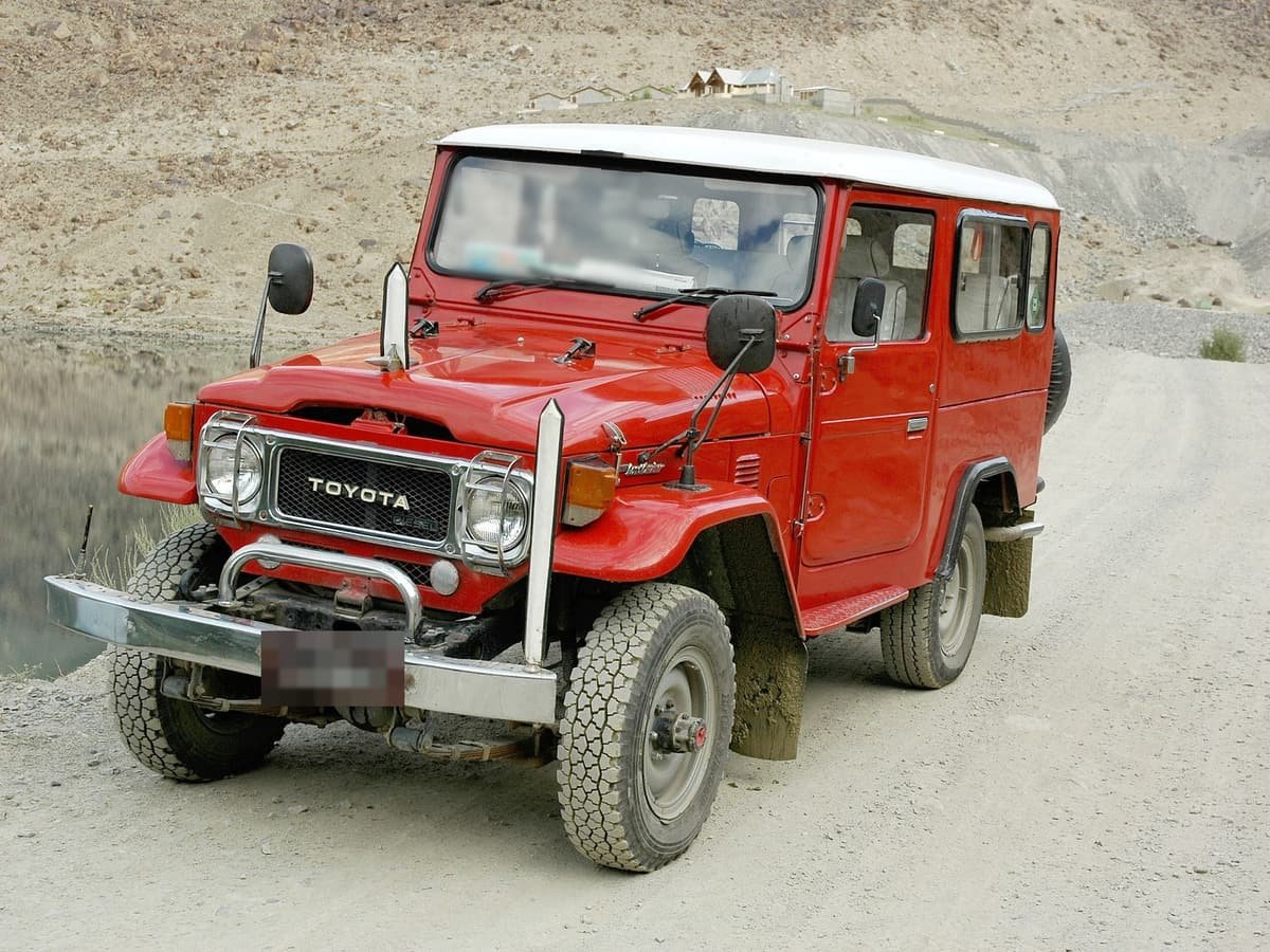 A red Land Cruiser jeep with a white roof on the gravel road to Deosai, a still lake and bare hills behind it